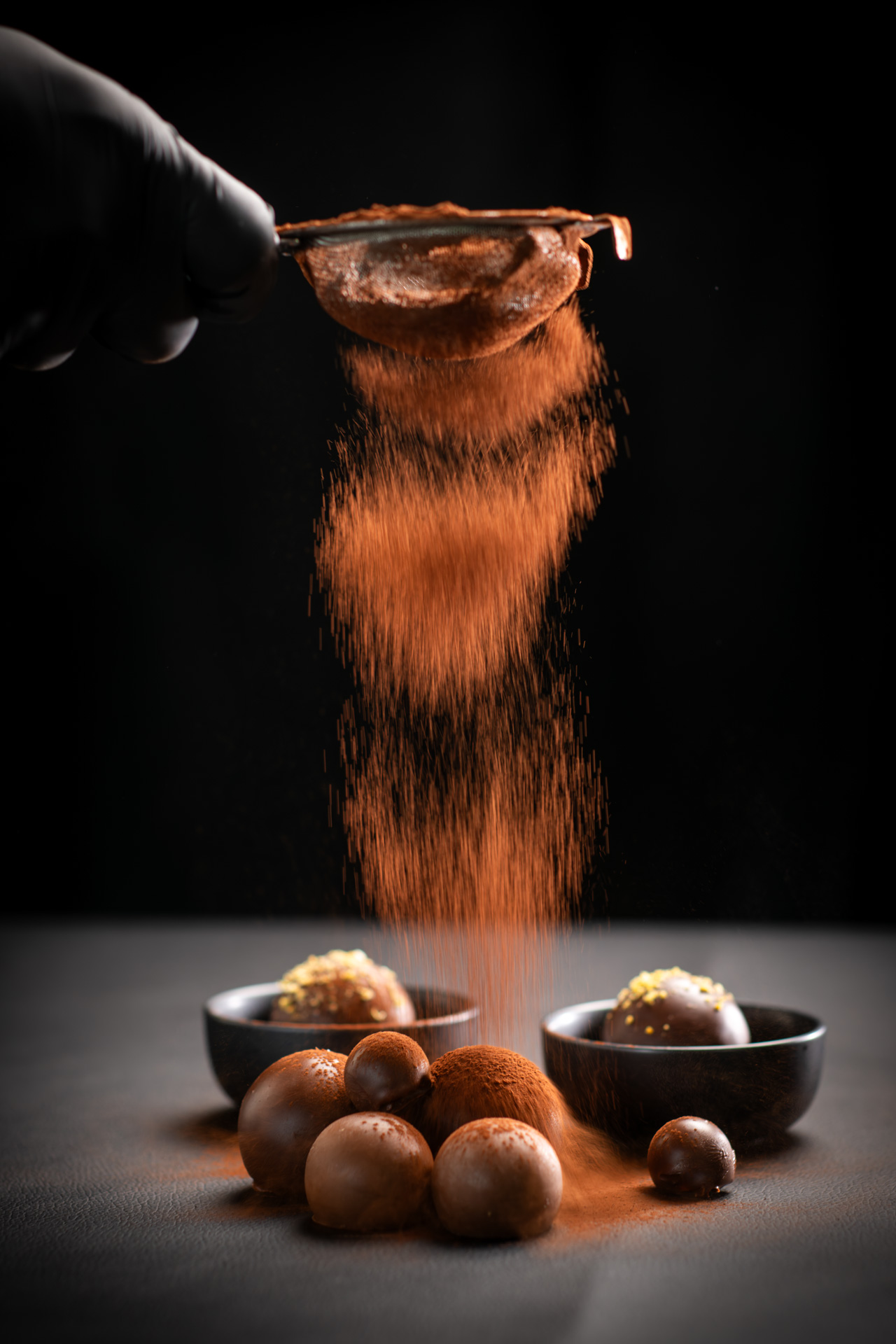 Behandschuhte Hand bestäubt Schokoladenpralinen mit Kakaopulver durch ein Sieb